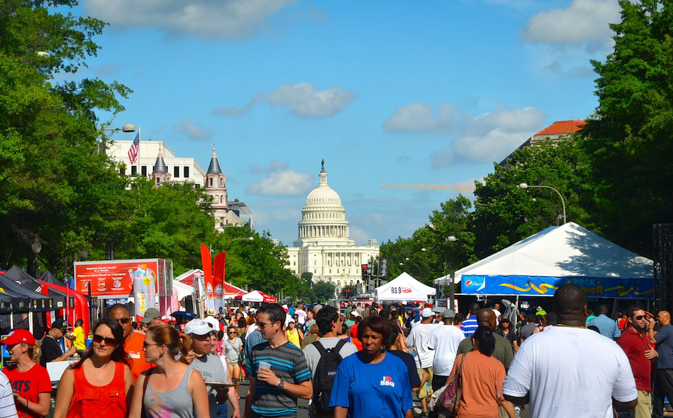 Festival Scene with Capitol Building: A busy festival scene on a sunny day with crowds of people walking between vendor tents and booths, the U.S. Capitol visible in the distance.