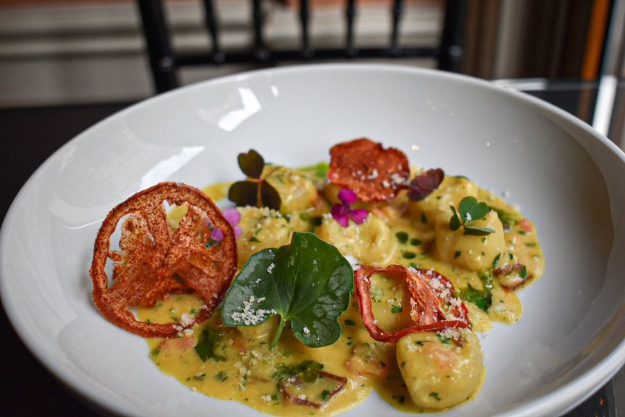 This image features a beautifully plated dish of pasta, garnished with fresh greens, thinly sliced tomatoes, and a creamy sauce. The presentation is enhanced with vibrant edible flowers, creating an appealing and elegant look.