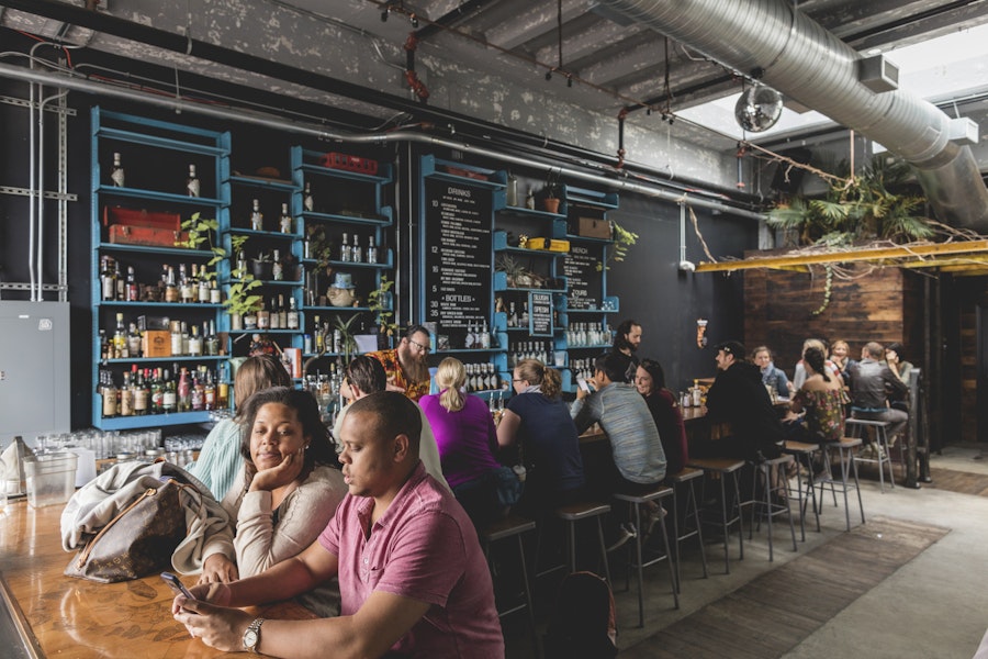 Patrons sitting and chatting at a cozy indoor bar with a well-stocked back wall of bottles.