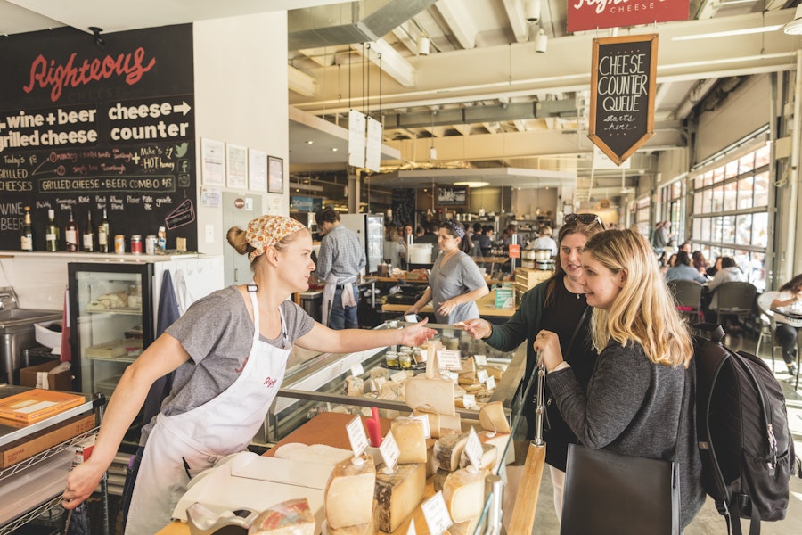 Customers interacting with a staff member at a cheese counter inside a busy market.