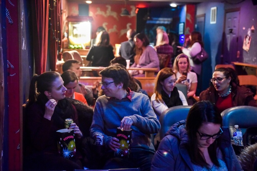 A group of people sitting in a cozy movie theater, eating popcorn and chatting before the film starts.