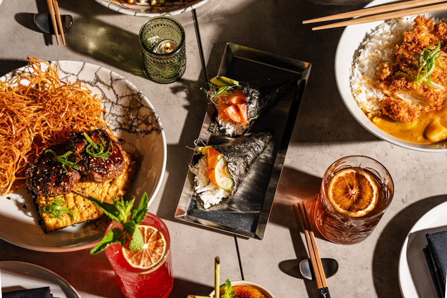 An overhead view of a diverse spread of Japanese dishes, featuring sushi rolls, rice bowls, and various appetizers, along with vibrant cocktails and meticulously plated dishes on a modern table setting.