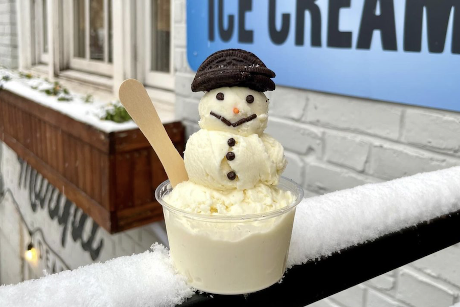 A small snowman made of vanilla ice cream scoops sits in a clear cup, with an Oreo cookie hat and chocolate chip eyes and buttons. The cup rests on a snowy ledge outside an ice cream shop, with a blue sign reading "ICE CREAM" visible in the background.