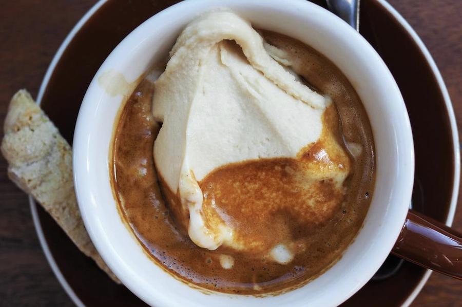 A close-up of a cup of affogato, featuring a scoop of gelato melting into a rich espresso. A biscotti rests on the side of the saucer.