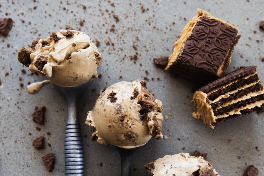 Two spoons of ice cream placed on a gray surface with chocolate chunks and layered chocolate wafers scattered around.