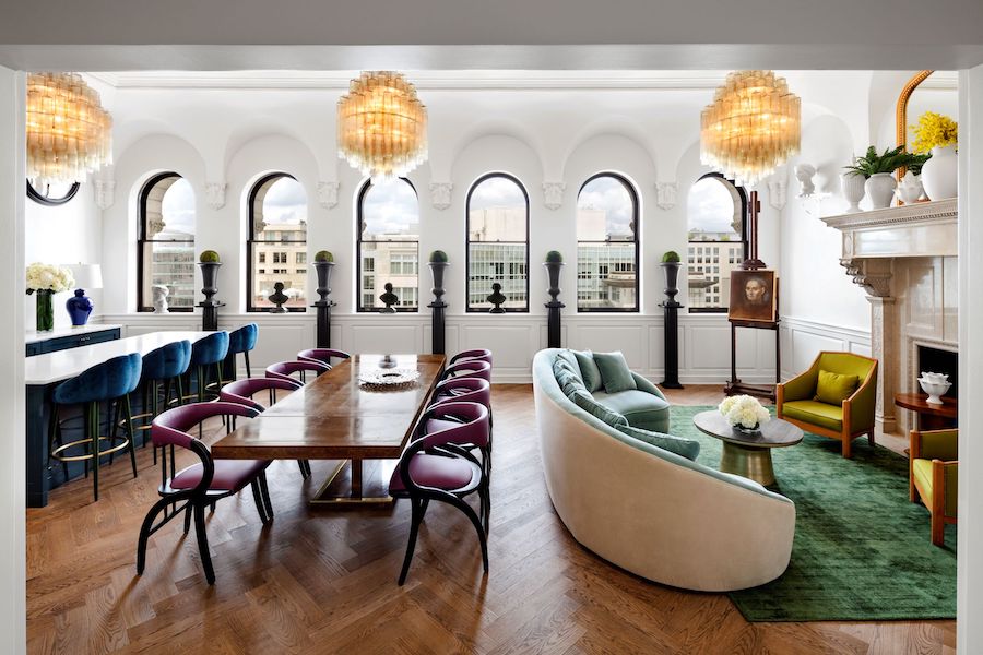 A stylish hotel lounge with a long wooden dining table, plush seating, arched windows offering city views, and elegant chandeliers hanging from the ceiling.