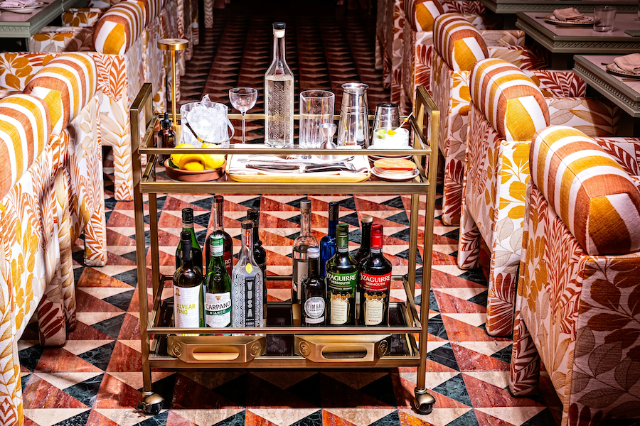 A stylish bar cart in a vibrant restaurant, stocked with various bottles of liquor, glassware, and a bowl of ice with citrus. The cart is set against a patterned geometric tile floor, and the seating features bold, floral and striped upholstery in warm tones.