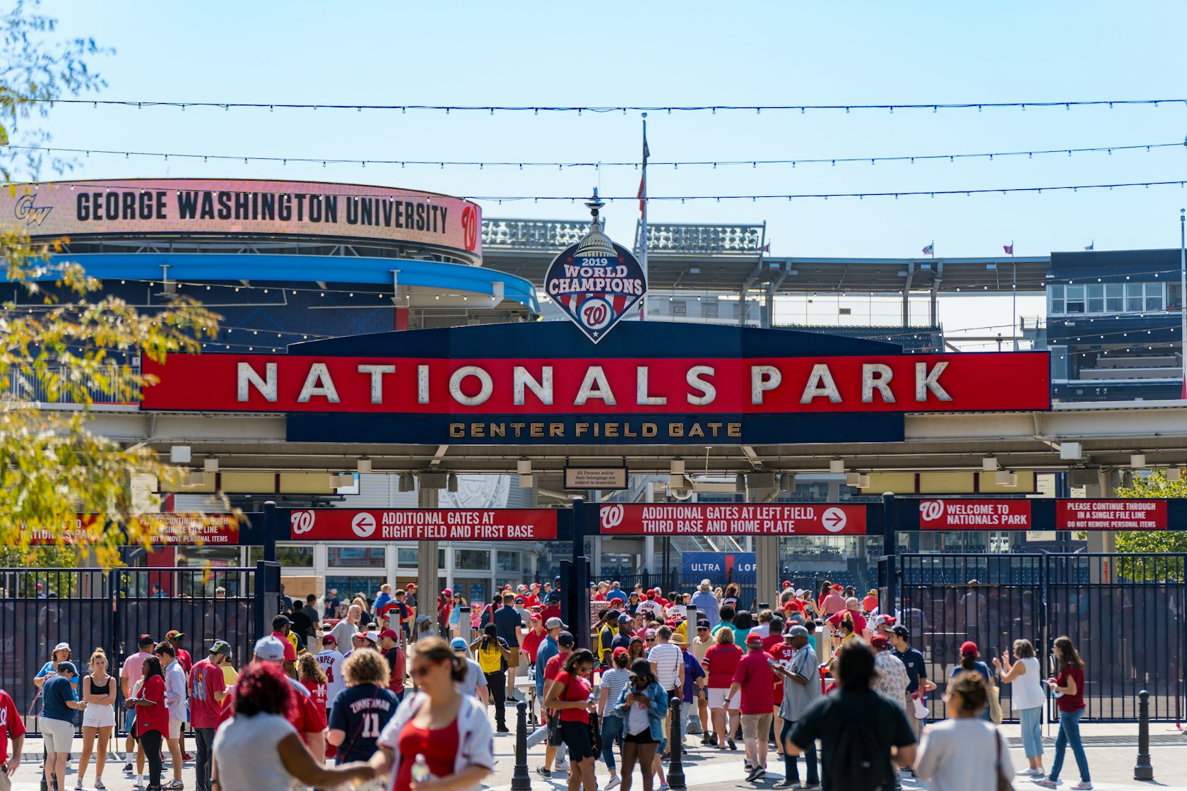 Crowds of fans dressed in red gather at the Center Field Gate of Nationals Park in Washington, DC, with a large "2019 World Champions" sign above the entrance.