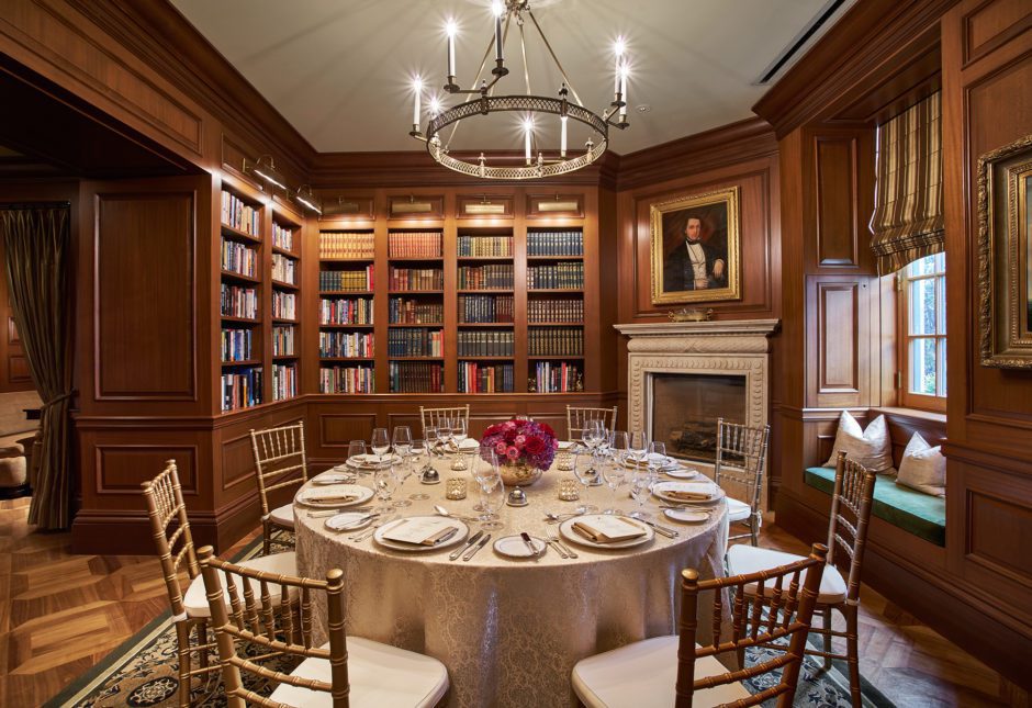 An intimate, wood-paneled library room at The Jefferson, featuring a round table set for a formal dinner, surrounded by bookshelves, a cozy fireplace, and elegant decor.