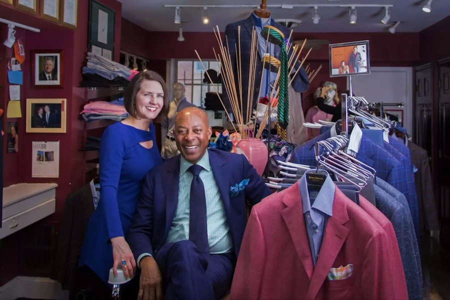 A well-dressed man and woman smile together in a tailored clothing store, surrounded by racks of colorful suits and neatly folded shirts, showcasing a stylish and welcoming atmosphere.