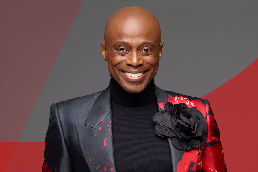 A portrait of the musician Kem, smiling and wearing a black and red suit with a large black flower detail on his shoulder, set against a stylish red and gray background.