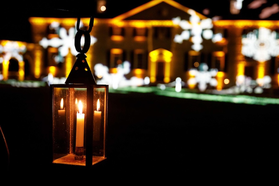 A lit candle lantern hangs in the foreground, with a large building illuminated in warm yellow lights and projected snowflake patterns in the background during an evening event.