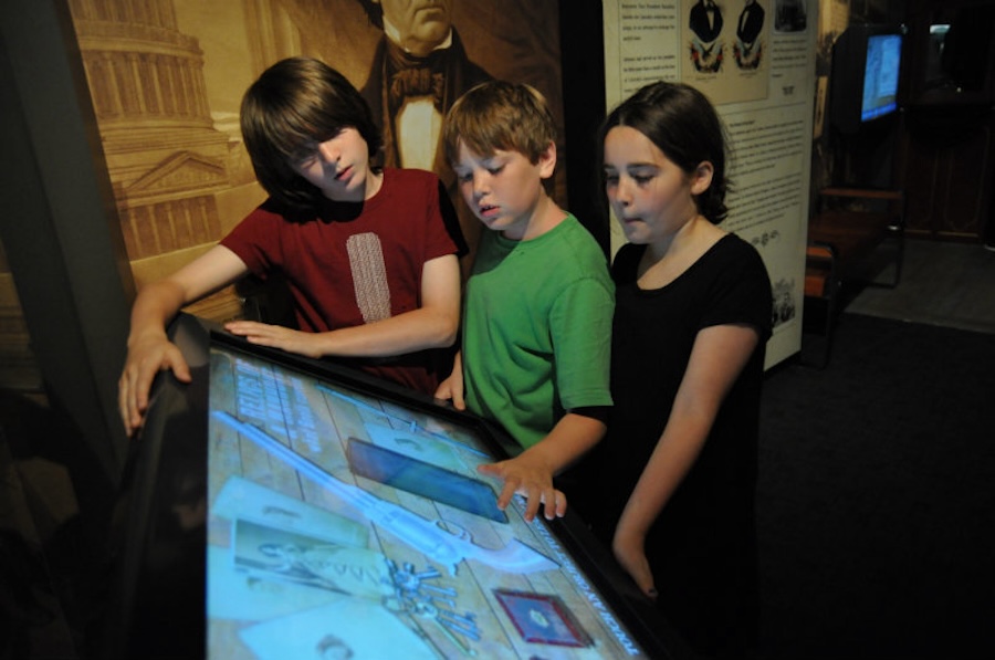 Three children interact with a touchscreen display at the Ford's Theatre, engaged in an educational activity related to history, with a large mural of Abraham Lincoln in the background.