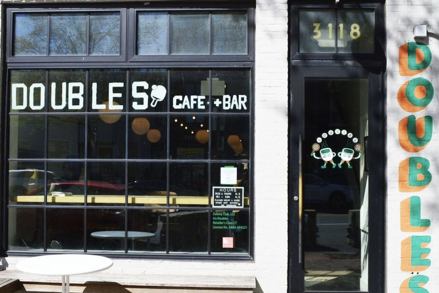 Exterior view of Doubles Cafe + Bar, showcasing large glass windows with the café's name in bold white letters. The black door displays the number "3118," and a small round table is set outside. Colorful graphics of ping pong paddles are part of the branding.