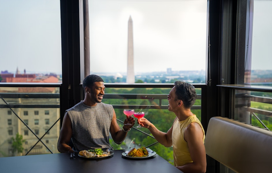 Two people enjoying a meal and toasting with drinks at a table with a view of the Washington Monument in the background, framed by large windows.