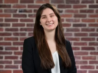 A woman with long brown hair, wearing a black blazer over a white blouse, stands in front of a brick wall.